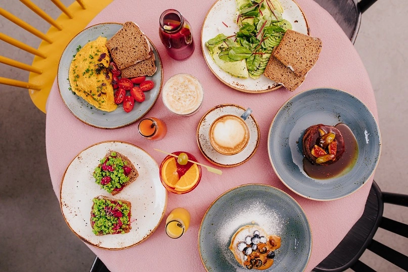 Brunch sobre una mesa rosa con tostadas, huevos, ensalada de hojas verdes, café con leche, jugo de naranja.